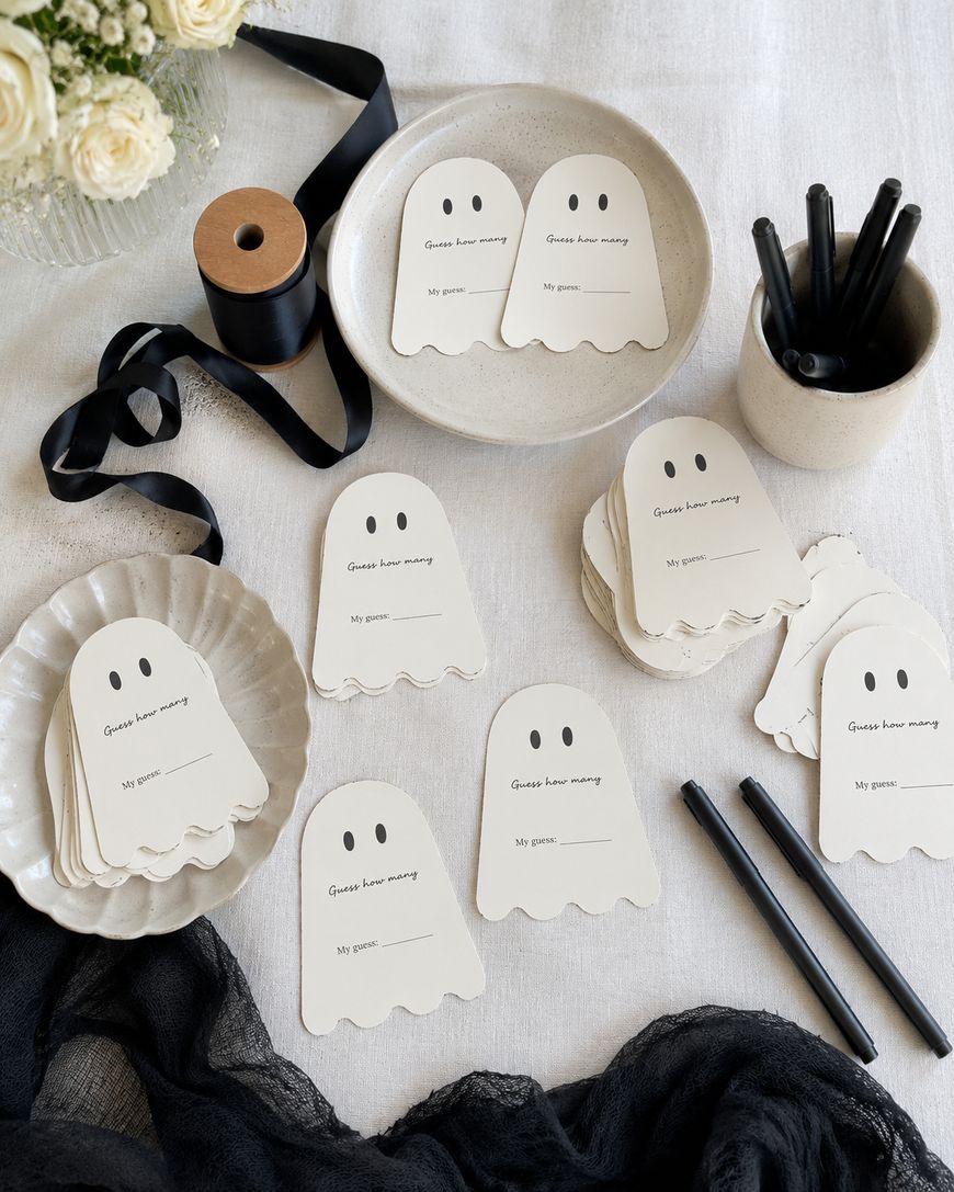 Stacked ghost-shaped cards labeled Guest name meaning, My guess, with black markers, ribbon, and white flowers on a light tablecloth. The scene is arranged neatly for a Halloween-themed activity.