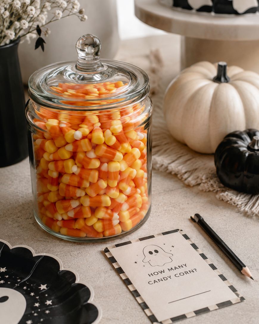 A glass jar filled with candy corn sits next to a notepad that says How many candy corn? surrounded by pumpkins and Halloween-themed decorations on a table.