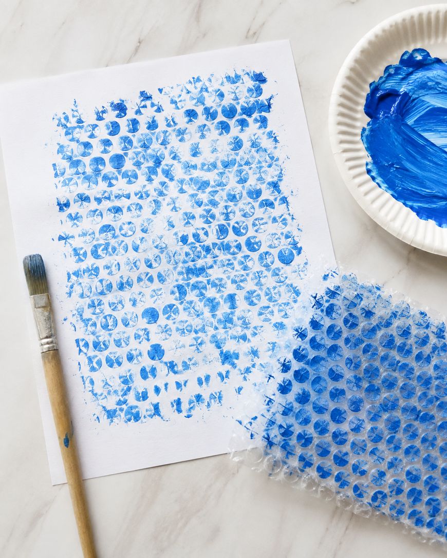 A sheet of white paper with blue paint dot patterns, a brush, a paper plate with blue paint, and a piece of painted bubble wrap on a white surface.
