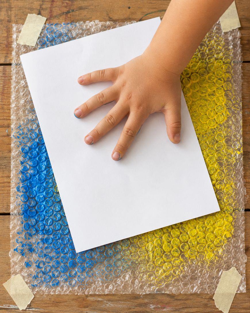 A childs hand presses white paper onto bubble wrap painted with blue and yellow on a wooden table, taped at the corners.