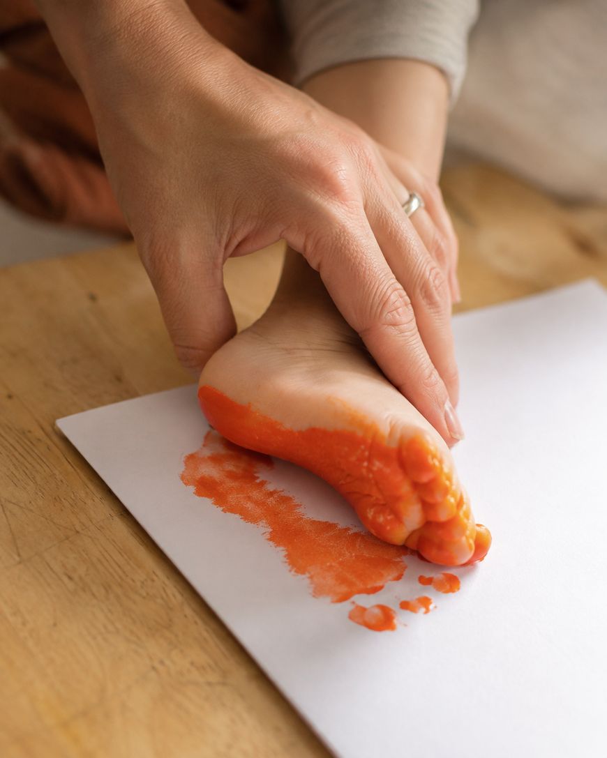 An adult presses a childs foot, covered in orange paint, onto a white sheet of paper to make a footprint print.