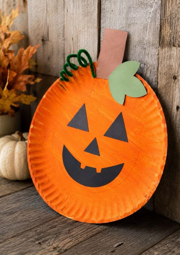 A paper plate painted orange is decorated as a jack-o-lantern with black paper facial features, a green leaf, a brown stem, and a green pipe cleaner vine. Its placed near autumn leaves and a white pumpkin.