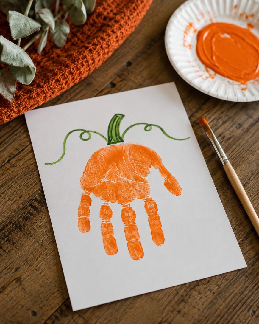 A handprint painted orange on white paper, resembling a pumpkin with green vines drawn on top. A paintbrush and a plate with orange paint are next to the paper on a wooden table.