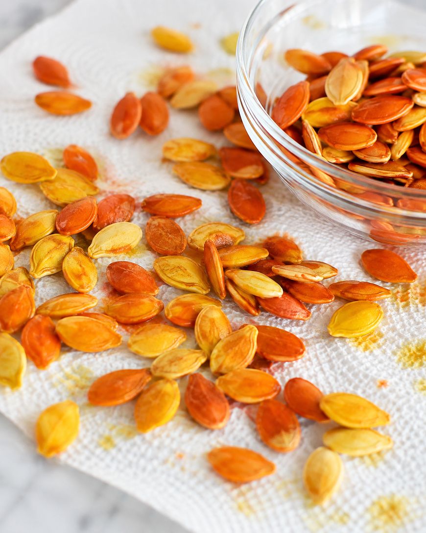 Pumpkin seeds, some orange and some yellow, are spread on a white paper towel with a glass bowl nearby, also holding seeds.