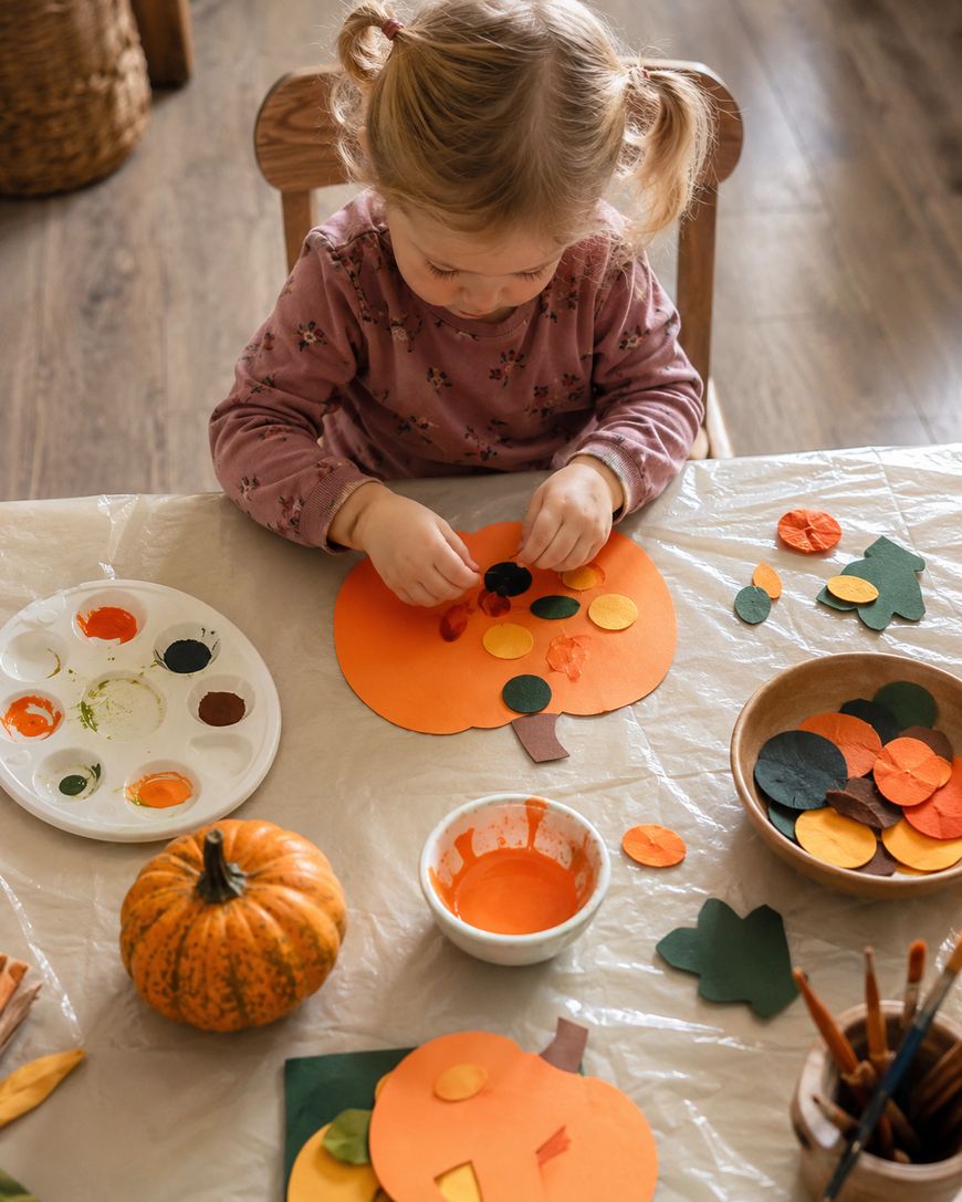 A young child sits at a table creating pumpkin crafts with colorful paper circles and paint. Art supplies, paint, a pumpkin, and more paper pieces are scattered around on the table covered with a plastic sheet.