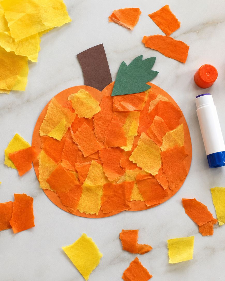 A pumpkin craft made from torn orange and yellow paper pieces, with a brown stem and green leaf, sits on a white surface alongside a glue stick and scattered paper scraps.
