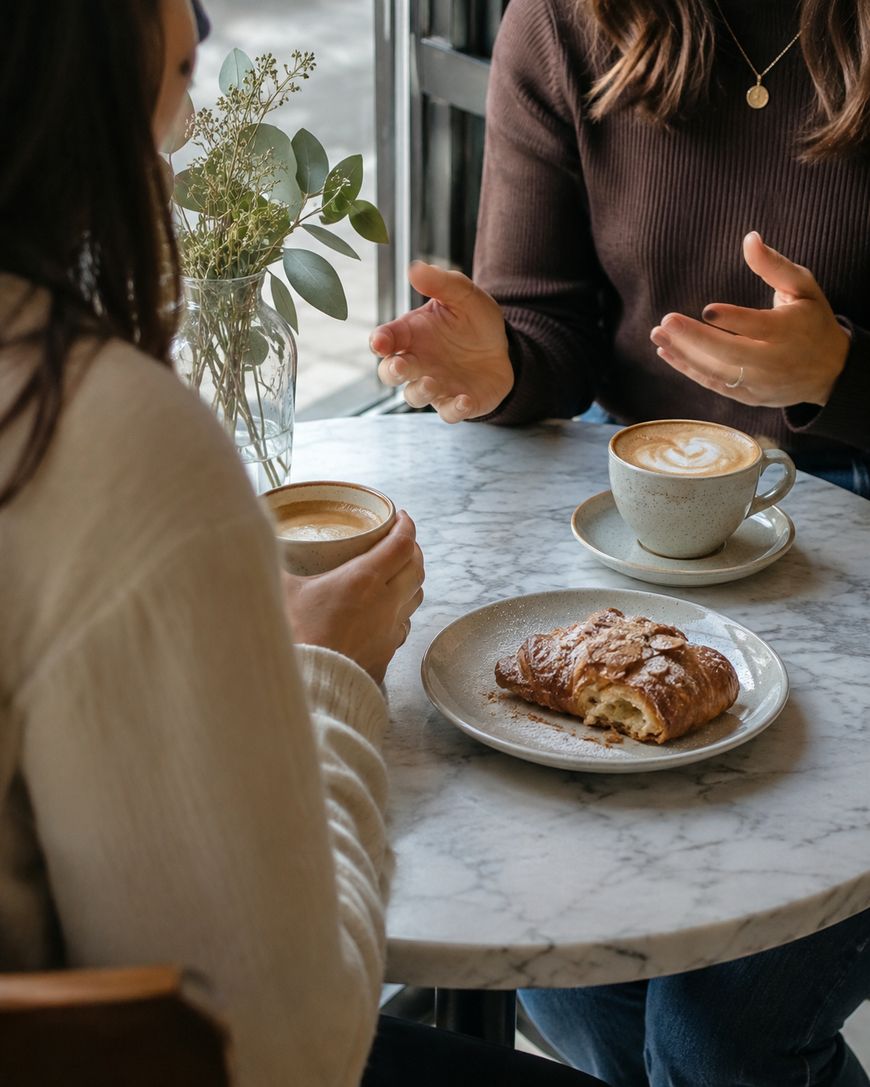 Two people sit at a marble table in a cafe, having coffee and a pastry. One person gestures with their hands, while the other holds a cup. A vase with flowers sits by the window.