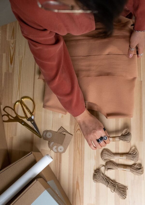 A person in a rust-colored outfit folds brown fabric on a wooden table, surrounded by scissors, twine, cardboard tags, and packaging materials—perfectly capturing the careful process that creative entrepreneurs use to prepare their handmade creations for shipping.