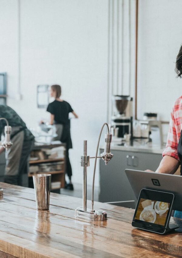 A person in a plaid shirt uses a tablet at a coffee shop counter, possibly exploring smart financial systems, while two others work in the background near coffee equipment.