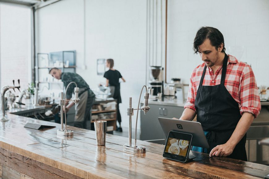 A barista in a red plaid shirt and black apron stands at a counter using a tablet register, showcasing efficient kitchen management; two other workers are busy in the background of a modern coffee shop.