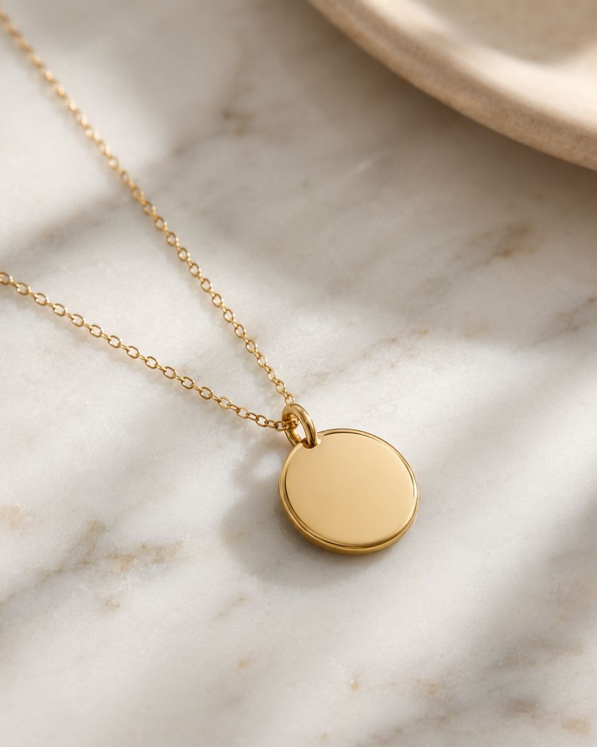 A gold round pendant necklace with a delicate chain rests on a marble surface, next to the edge of a beige ceramic dish.