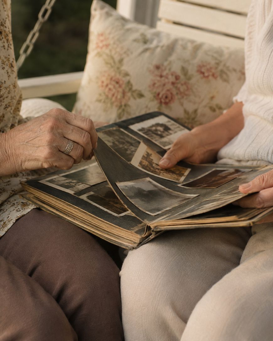 Two people sit closely together, flipping through an old photo album filled with black-and-white pictures, suggesting shared memories and nostalgia. Only their hands and part of their laps are visible.