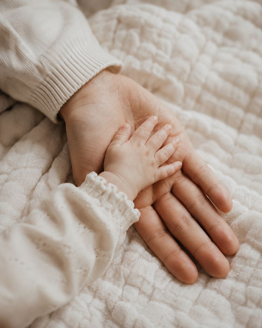 An adult hand resting on a soft, quilted blanket with a babys hand gently placed on top of it, both wearing light-colored long sleeves.