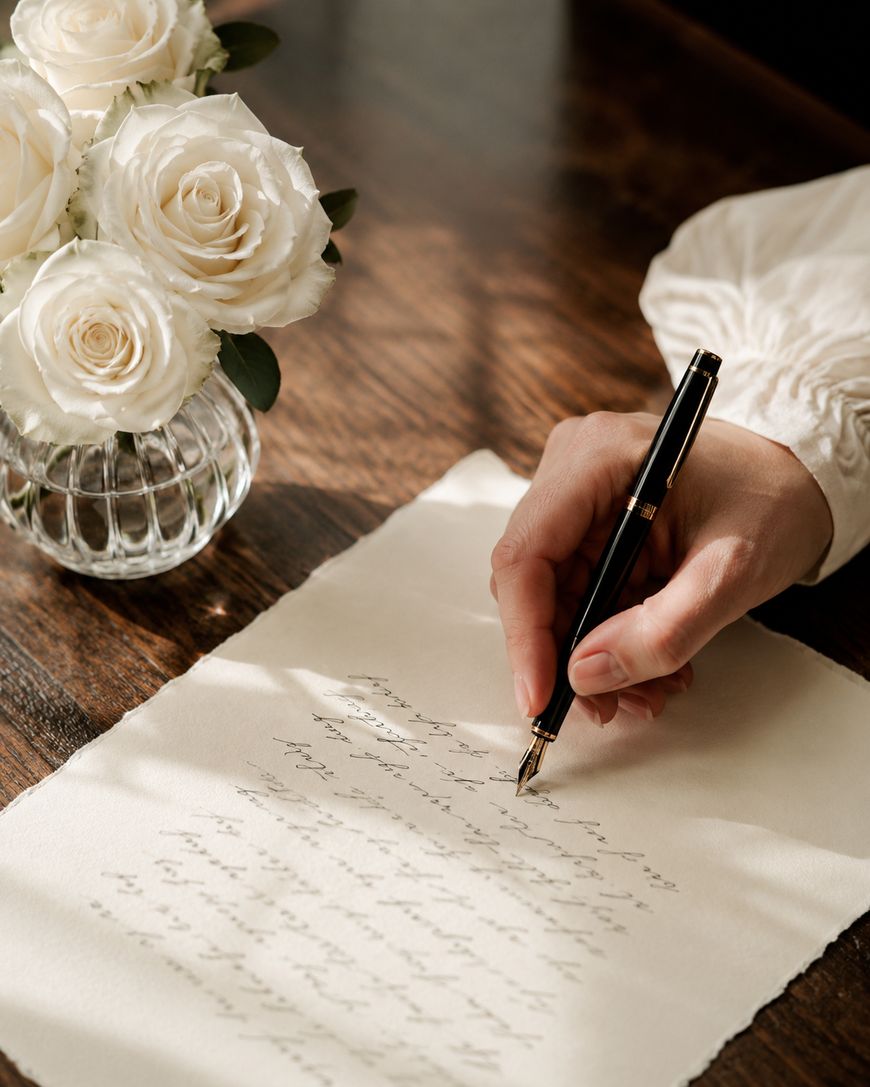 A hand writes a letter with a fountain pen on cream paper. A vase with white roses sits on the wooden table nearby. Sunlight streams in, casting soft shadows.