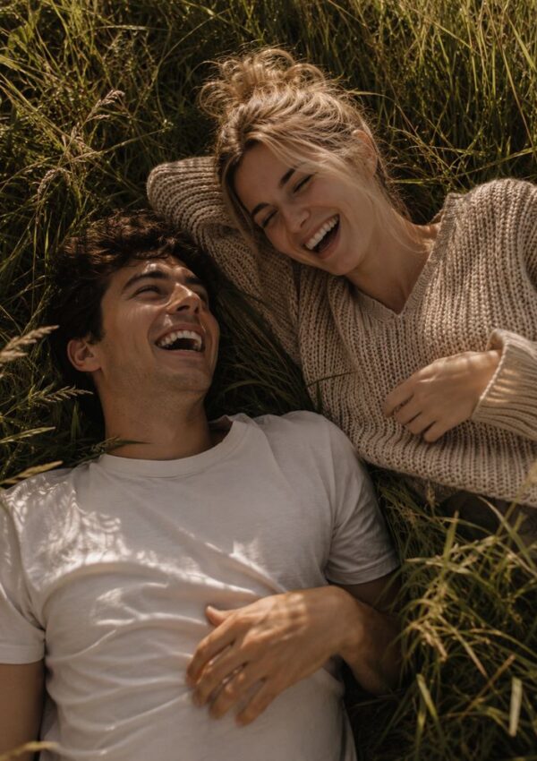 A young man and woman lie on grass, smiling and laughing together. The woman wears a beige sweater; the man wears a white t-shirt. Sunlight highlights their faces, creating a warm, joyful atmosphere.