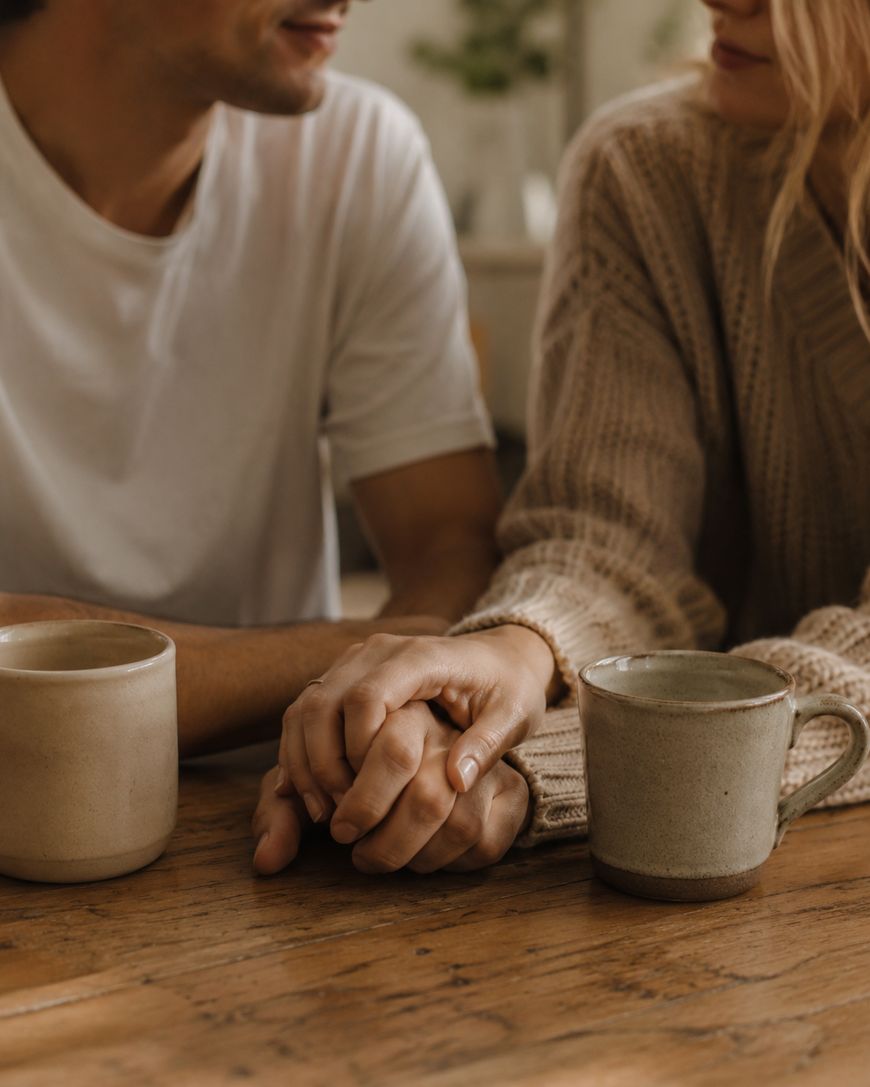 Two people sit at a wooden table, holding hands. Each has a light-colored mug in front of them. One person wears a white shirt, the other a beige sweater. The scene feels warm and intimate.