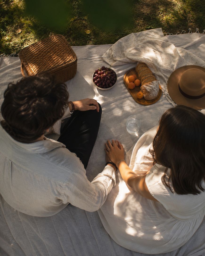 A couple sits on a blanket outdoors, holding hands. A picnic basket, bread, grapes, apricots, a hat, and a glass of water are arranged beside them on the blanket under dappled sunlight.