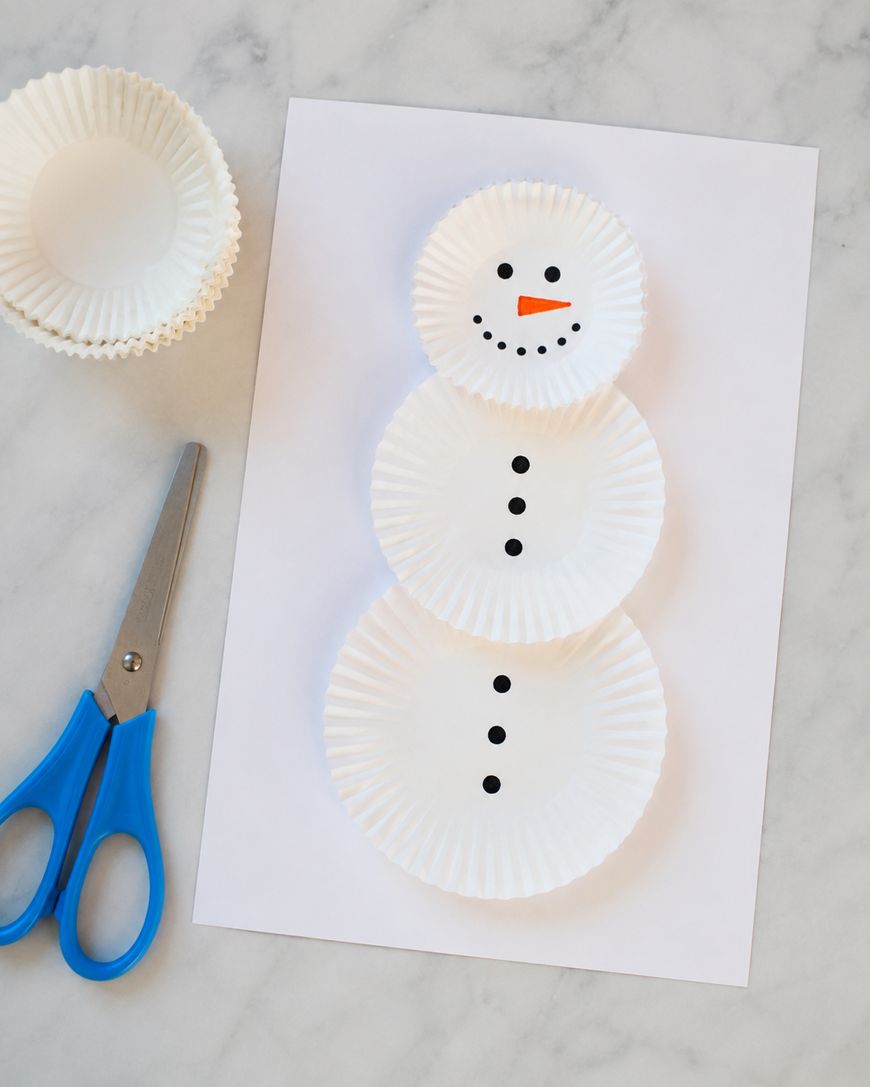 Three white paper cupcake liners are arranged on a sheet of paper to form a snowman, with marker-drawn eyes, buttons, smile, and an orange carrot nose. Blue scissors and extra liners are beside the artwork.