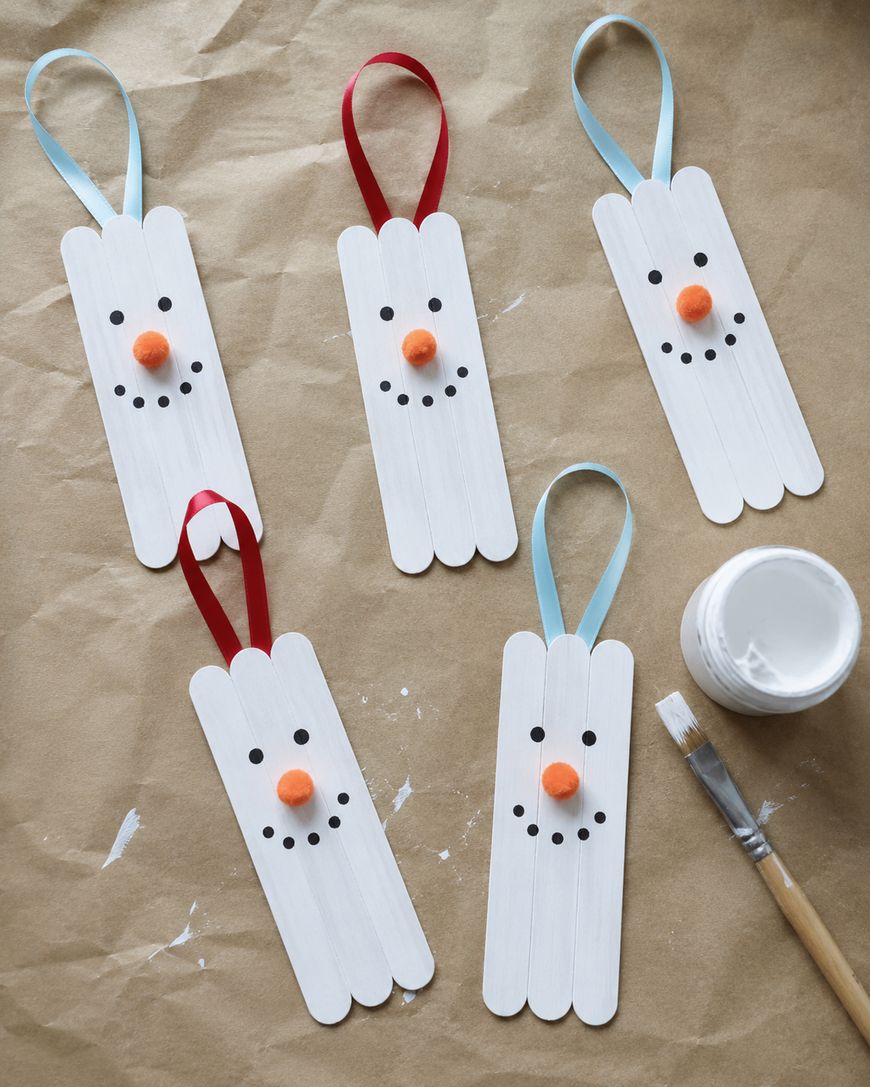 Five snowman ornaments made from painted craft sticks, each with a ribbon loop, black dot eyes and mouths, and orange noses, are arranged on brown paper next to a paintbrush and an open white paint jar.