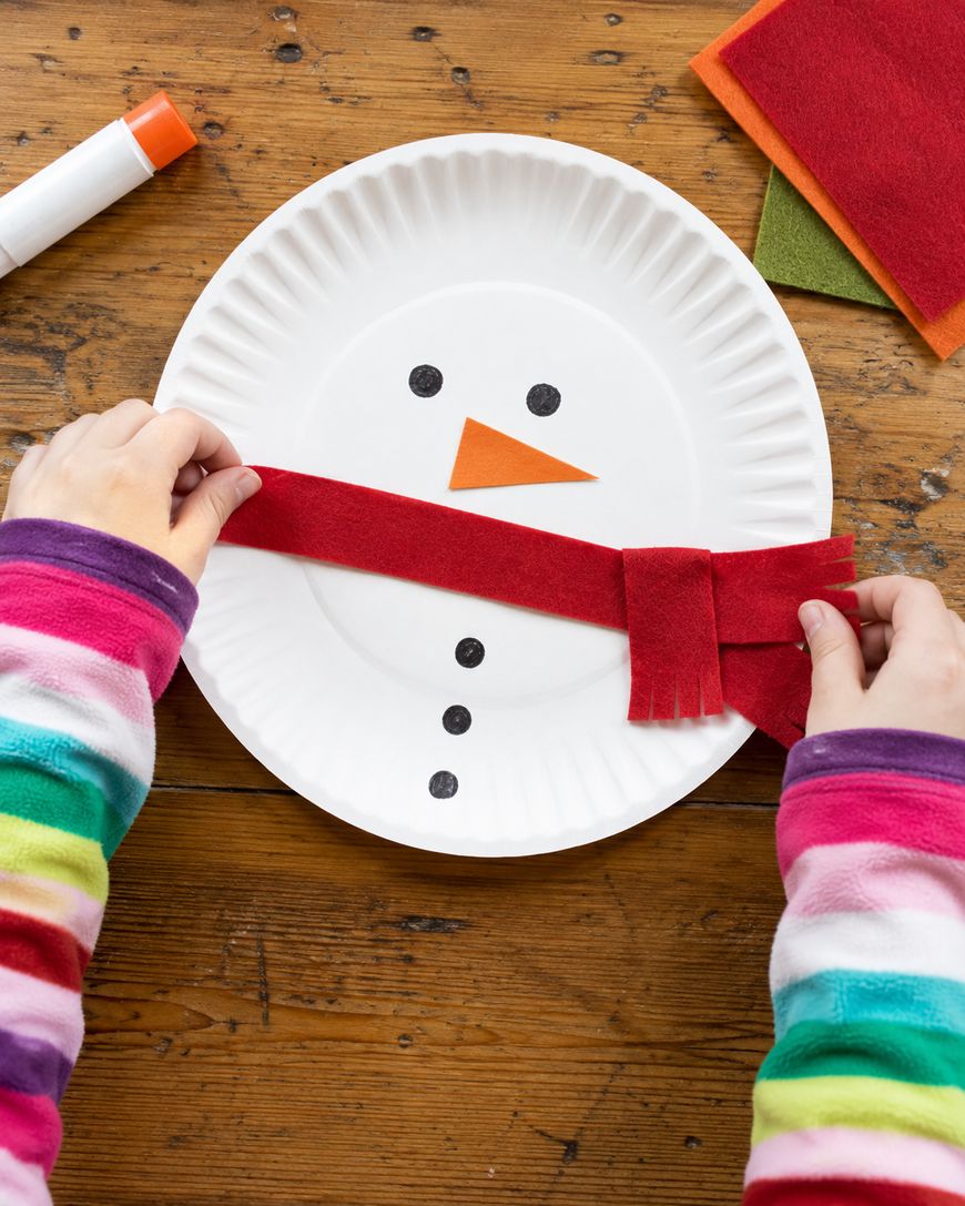 A child, wearing a colorful striped sweater, crafts a snowman face on a paper plate, adding a red felt scarf. The snowman has a carrot nose, black eyes, and buttons. Glue stick and felt sheets are on the wooden table.