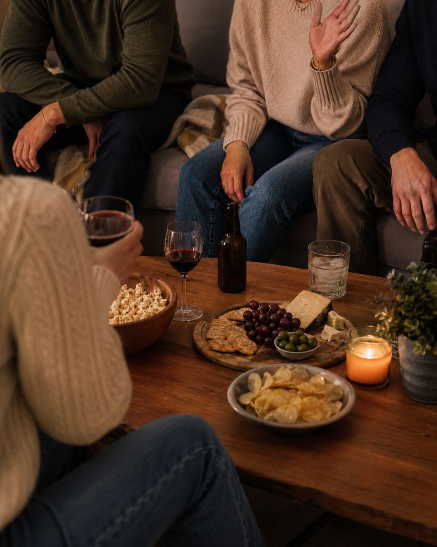 Three people sit on a couch and one in front, enjoying snacks, wine, and beer on a coffee table with chips, popcorn, cheese, grapes, crackers, olives, and a lit candle. Only their torsos and hands are visible.