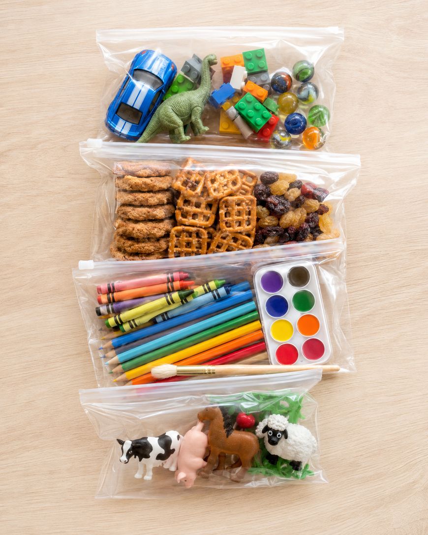 Four clear zip bags on a wooden surface, each containing different items: toy car, dinosaur, Lego bricks, marbles; snacks; crayons, pencils, watercolor paints; and toy farm animals with plastic grass.