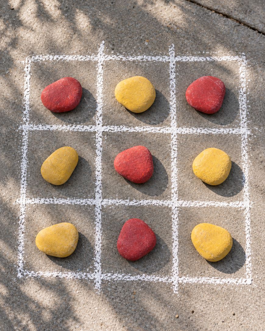Tic-tac-toe grid drawn in white chalk on concrete, filled with yellow and red painted rocks.