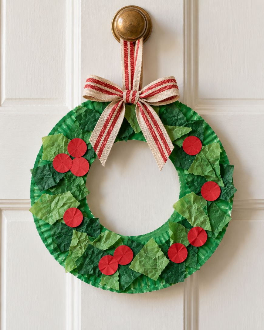 A homemade wreath made from a green paper plate, decorated with green tissue paper squares and red paper circles, hanging from a door by a red and white striped ribbon.