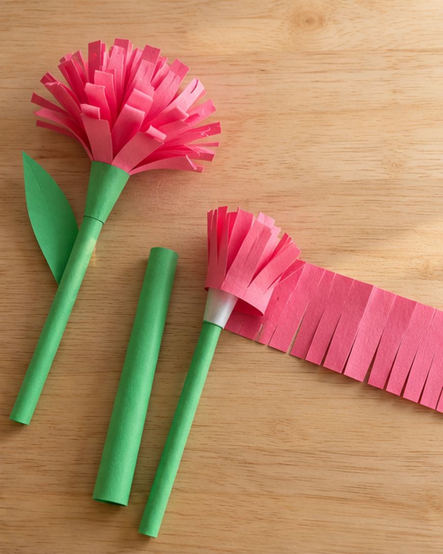 Two paper carnations on a wooden surface, one finished and one in progress with a strip of pink fringe being attached to a green stem.