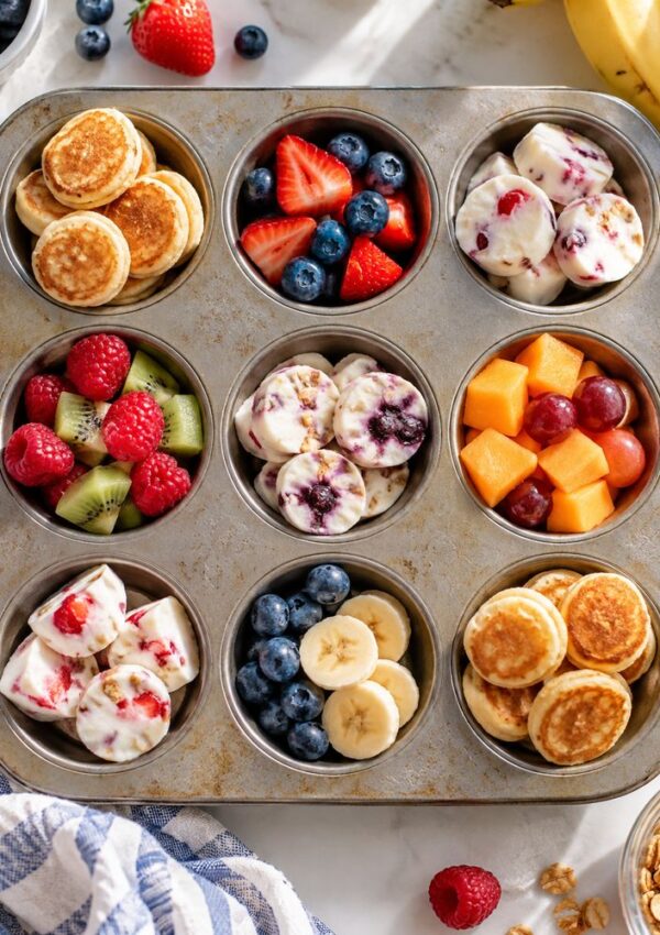 A muffin tin filled with nine servings of breakfast snacks, including mini pancakes, yogurt bites, and bowls of fresh fruit like strawberries, blueberries, kiwi, banana slices, and melon.