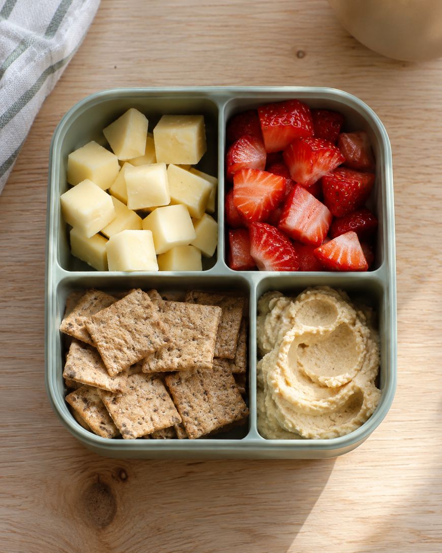 A square snack container divided into four sections containing cubed cheese, sliced strawberries, whole grain crackers, and hummus.