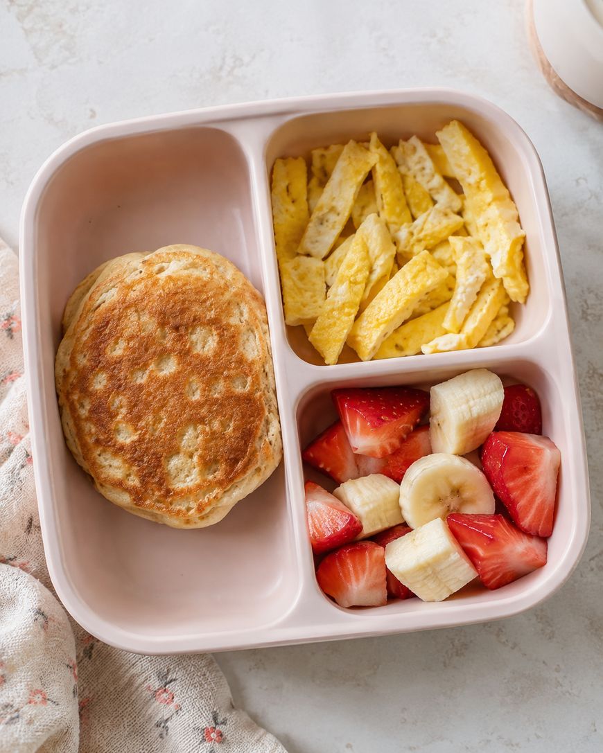 A divided tray containing a pancake, strips of cooked egg, and a mix of chopped strawberries and bananas.